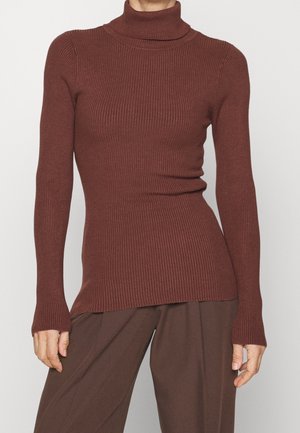 Femme portant un pull à col roulé côtelé marron ajusté et un pantalon marron foncé ample sur un fond uni.