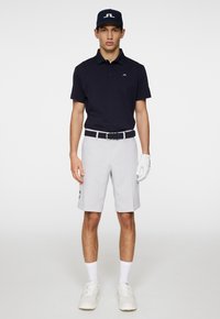Mörkblå polo skjorta, ljusgrå shorts och vita sneakers. Svart bälte med mönstrad detalj. Modellen håller en golfhandske och bär en matchande keps.