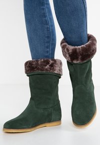 Jambes dans un jean bleu portant des bottes en suède vert foncé avec des poignets en fourrure synthétique brune repliés et des semelles plates marron clair sur un sol blanc.