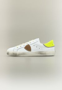 Basket blanche basse avec semelle usée, accent jaune au talon, patch logo beige sur le côté et lacets blancs sur fond neutre.