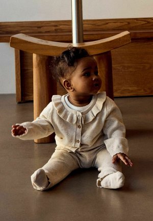 Bambino piccolo con capelli scuri e ricci, vestito con un completo lavorato a maglia color crema, seduto sul pavimento vicino a una sedia di legno in una stanza con pannelli di legno.