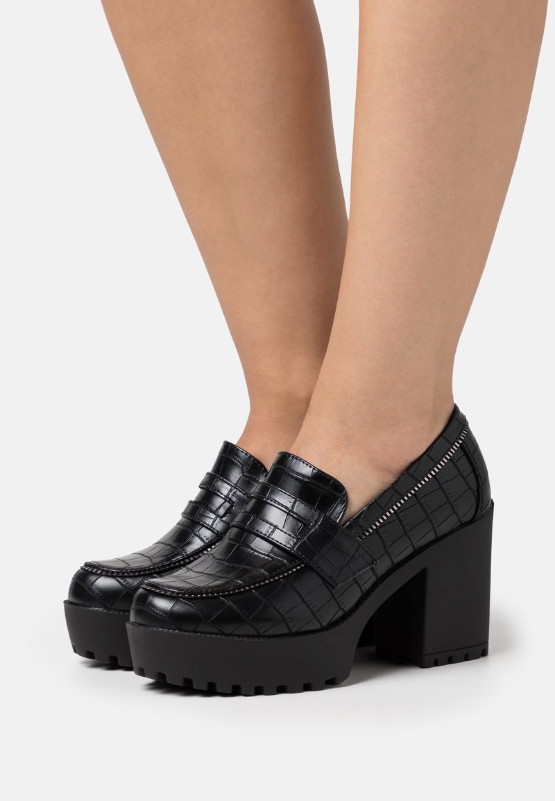 Pieds portant des mocassins à plateforme noirs embossés effet croco, avec des talons hauts épais et des semelles rainurées sur un fond clair.