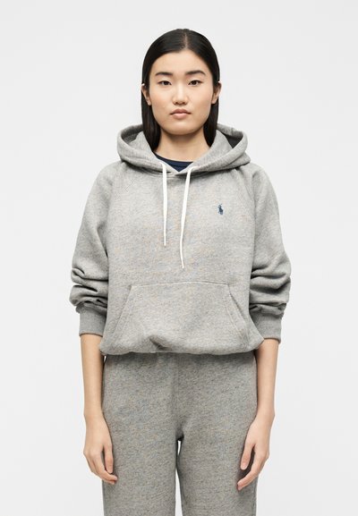 Νεαρή γυναίκα με ίσια σκουρόχρωμα μαλλιά, ντυμένη με γκρι φούτερ με άσπρα κορδόνια και ταιριαστό γκρι φόρμα, στέκεται μπροστά από ένα απλό φόντο.