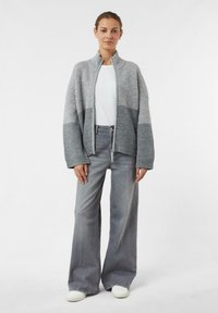 Grauer gestrickter Cardigan mit hohem Kragen und offener Front, kombiniert mit grauen weiten Jeans und weißen Sneakers. Glatte Textur und lässige Passform.