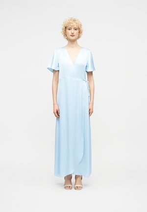 Γυναίκα με κοντά σγουρά ξανθά μαλλιά που φοράει ένα ανοιχτό μπλε φόρεμα τύπου wrap μέχρι τον αστράγαλο και μπεζ πέδιλα με ανοιχτές μύτες, στέκεται μπροστά σε λευκό φόντο.