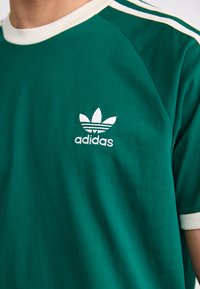 Zöld pamut póló fehér csíkkal a galléron és a vállon; fehér hímzett Adidas logóval rendelkezik, amely három levélből áll.