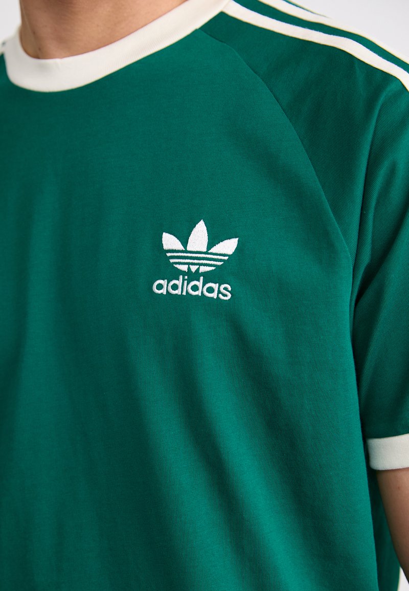 Camiseta de algodón verde con cuello de canalé blanco y rayas en los hombros; presenta un logotipo bordado de Adidas en blanco con un diseño de tres hojas.