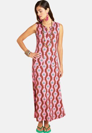 Woman wearing sleeveless pink patterned maxi dress with green sandals, colorful beaded necklace, pink earrings, and bracelets, standing with hand on hip.