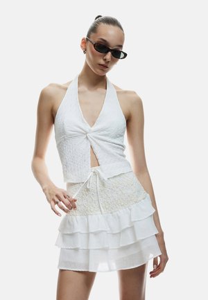 Witte halter-top van textuurstof met een geknoopte voorkant, gecombineerd met een gelaagde minirok met kantaccenten. Model draagt een zonnebril.