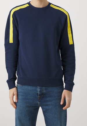 Man draagt een marineblauwe trui met gele strepen en de tekst "EA7 Emporio Armani" op de schouders, gecombineerd met een blauwe spijkerbroek.