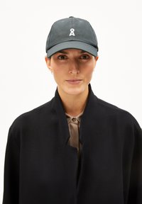 Vrouw met een donker petje met een wit symbool, een beige overhemd met knopen en een zwart open jasje tegen een witte achtergrond.