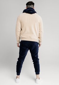 Felpa in corduroy beige con colletto a coste blu navy, abbinata a joggers in corduroy blu navy; presenta polsini elastici e sneakers bianche.