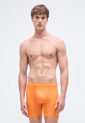 Orange cotton boxer briefs with an elastic waistband, featuring a smooth texture and fitted design, worn by a model with a neutral expression.