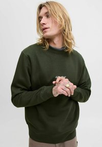 Plavi muškarac s tetovažama i prstenom nosi tamnozelenu duksericu JJStudios, gleda u stranu s rukama preklopljenim na pozadini jednostavnog svijetlog tona.