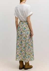 Weißes Kurzarmhemd, kombiniert mit einem Blumen-Maxirock, der rosa, blaue und gelbe Muster aufweist. Braune Knöchelschäfte runden den Look ab.