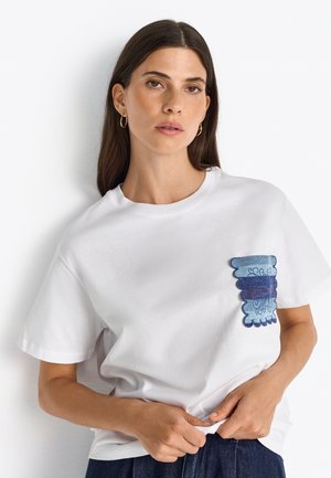T-shirt en coton blanc avec une poche en denim, présentant des nuances de bleu et des coutures décoratives. Manches courtes, coupe décontractée et col rond.