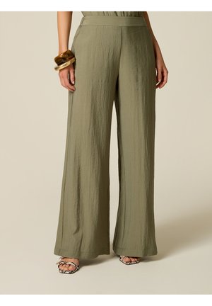 Femme portant un pantalon large vert olive avec des sandales ouvertes à imprimé serpent et des bracelets épais au poignet gauche sur un fond neutre.