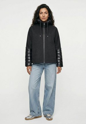 Frau trägt eine schwarze Kapuzenjacke mit gesteppte Ärmel und hellblaue Jeans mit weitem Bein, steht vor einem schlichten hellen Hintergrund.