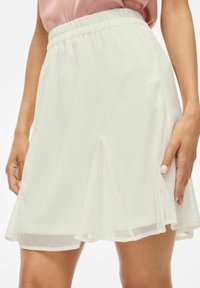 Jupe beige clair avec une taille élastique, fabriquée en tissu transparent, dotée d'un design évasé et d'une fente subtile à l'ourlet.