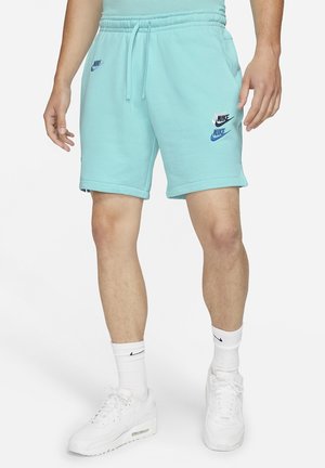 Homme portant un short Nike bleu clair avec cordon de serrage et trois logos Nike, des chaussettes blanches Nike et des baskets blanches, debout sur un fond blanc.