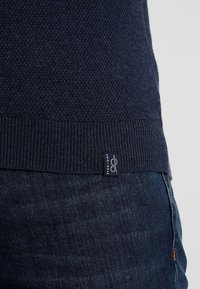 Marineblå stickad tröja med strukturerat mönster, ribbade muddarna och midjan. Innehåller en liten etikett i botten med "Pier One"-logotyp.