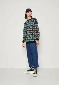 Jovem de boné preto, suéter padrão azul e laranja, jeans largos azuis e tênis alto pretos está encostado contra um fundo liso.