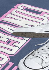 Camiseta azul con gran texto gráfico en blanco y rosa, con una ilustración impresa de una zapatilla blanca y un diseño detallado de la suela.