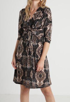 Vrouw die een knielange jurk draagt met zwarte, beige en bruine paisley-patronen en driekwartmouwen, voorzien van een knoopdetail aan de voorkant.