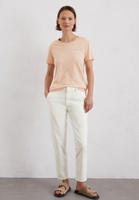 Light peach short-sleeve t-shirt with a round neck and subtle text detail, paired with white straight-leg trousers and beige sandals.