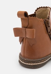 Brązowy skórzany but za kostkę z falistą górną krawędzią, tylną pętelką i detalem w postaci kokardy. Posiada teksturowaną podeszwę z białymi akcentami w postaci stębnowania.