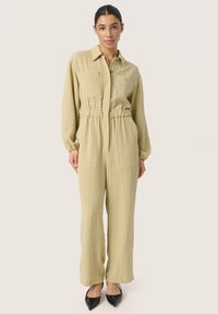 Lys khaki utility jumpsuit med lange ærmer, to forlommer og elastisk talje. Båret med spidse sorte hæle. Glat tekstur.