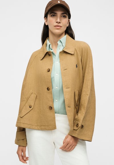 Γυναίκα που φοράει μπεζ σακάκι με κουμπιά πάνω από ένα ανοιχτό μπλε πουκάμισο, άσπρο παντελόνι και καφέ μπέιζμπολ καπέλο, στέκεται μπροστά σε μια απλή φόντο.