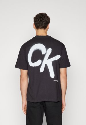 Czarny bawełniany T-shirt z dużym białym logo "CK" w stylu spray'u na plecach oraz małą metką marki na dole po prawej stronie.