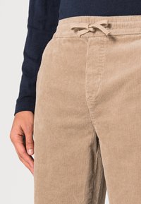 Beige corduroy broek met verticale ribbels, voorzien van een elastische tailleband met een gescherpte koord. Close-up van een hand die op de stof rust.