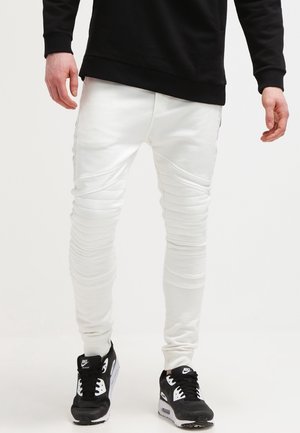 Mężczyzna w białych spodniach w prążki, czarnej bluzie i czarno-białych trampkach Nike stoi na białym tle.