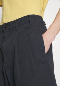Black pleated trousers with side pockets, featuring a button closure and a smooth fabric texture, paired with a yellow shirt.