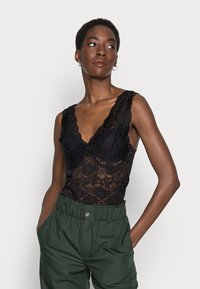 Top in pizzo nero con fantasie floreali, scollatura a V e spalline larghe, abbinato a pantaloni verdi a vita alta con dettaglio a bottoni.