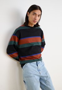 Sudadera negra con rayas horizontales en verde, marrón y azul, combinada con unos vaqueros de mezclilla azul claro. Corte relajado, tejido suave.