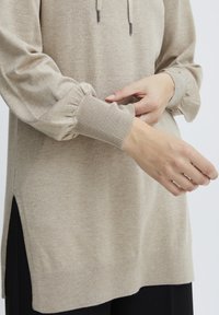 Sweat à capuche en maille beige, avec des manches froncées et des poignets côtelés. Fentes latérales notables et finition douce et texturée.