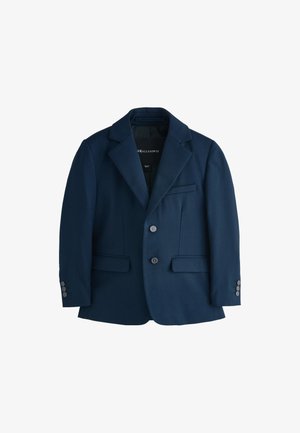 Ragazzo con i capelli corti che indossa un completo blu scuro, camicia marrone e sneakers bianche, in piedi con una mano in tasca e guarda di lato.