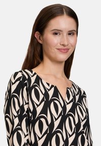 Schwarz und cremefarbige Bluse mit V-Ausschnitt, die ein abstraktes Design aus Formen aufweist, gefertigt aus einem leichten Stoff mit glatter Textur.