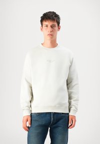 Young man wearing a light beige sweatshirt with "Replay" logo and blue jeans, standing against a plain white background.