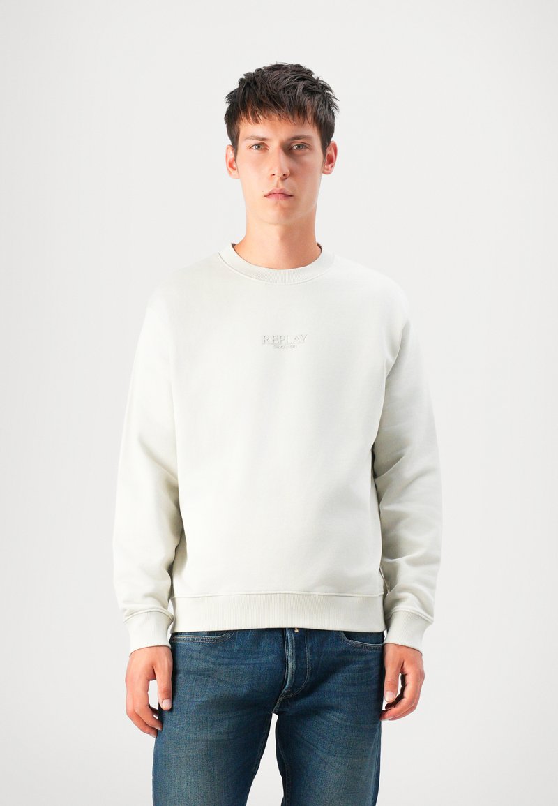 Young man wearing a light beige sweatshirt with "Replay" logo and blue jeans, standing against a plain white background.
