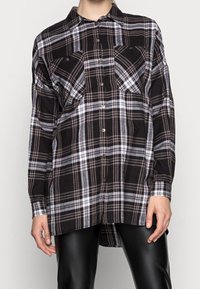 Camicia a scacchi bianco e nero con maniche lunghe, apertura con bottoni sul davanti e due tasche sul petto. Il tessuto sembra texturizzato e morbido.