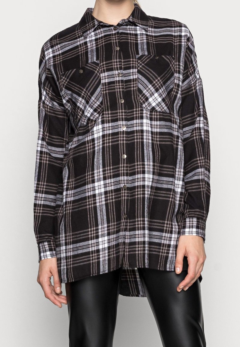 Camicia a scacchi bianco e nero con maniche lunghe, apertura con bottoni sul davanti e due tasche sul petto. Il tessuto sembra texturizzato e morbido.