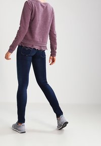 Sudadera púrpura con dobladillo festoneado, combinada con jeans ajustados azul oscuro y zapatillas deportivas grises. Enfocándose en un diseño casual y entallado.