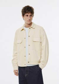 Lys beige jakke lavet af blødt stof, med knaplukning, to forlommer og en afslappet pasform. Båret over en lys sweater.