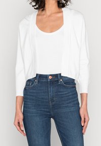 Biały, lekki kardigan z długimi rękawami, otwarty z przodu, o luźnym kroju, noszony na prostym białym topie na ramiączkach i ciemnoniebieskich dżinsach.