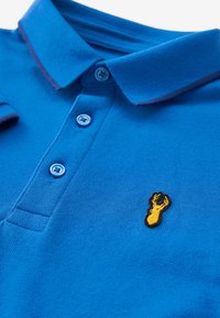 Polo de color azul con una tela texturizada, que presenta un logo de langosta bordado en amarillo en el lado izquierdo y dos botones azules en el cuello.