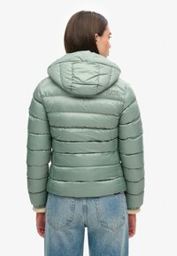 Chaqueta verde acolchada con capucha, que presenta un diseño acolchado horizontal, textura suave y detalle del logo en la parte posterior. Combinada con unos jeans azul claro.